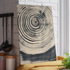 May include: Rectangular cutting board with a black and white abstract design resembling tree rings. The artwork features concentric circles and a dark, brushstroke-style base. The board is displayed near a window and kitchen utensils.