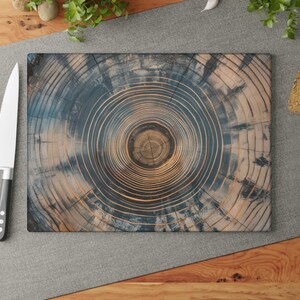 May include: A rectangular wooden cutting board with a tree ring design in shades of blue and brown. A large knife with a silver blade and black handle rests beside the board, along with red tomatoes. The board is surrounded by food items and greenery.