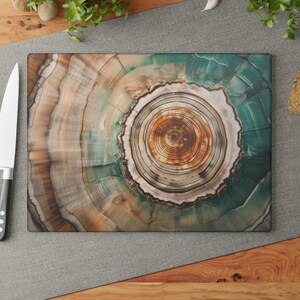 May include: A rectangular cutting board with a tree ring design in shades of brown, green, and white. A stainless steel knife with a black handle and three red tomatoes are on the left. Bread and garlic are on the right.