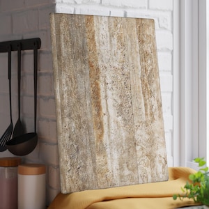 May include: A rectangular chopping board with a natural wood grain pattern. The board is beige and brown, with vertical lines and a textured surface. It is leaning against a white brick wall near a window.