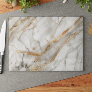 May include: Rectangular white marble cutting board with gold veining. A stainless steel knife with a black handle and three red tomatoes are on the left. Slices of bread, garlic, and herbs are in the background.