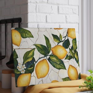 May include: Rectangular cutting board with a lemon and leaf design. The board features yellow lemons, green leaves, and white flowers against a cracked white background. The board is resting on a yellow cloth.