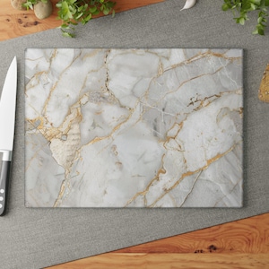 May include: Rectangular cutting board with a marble design featuring gray and gold veining. A stainless steel knife with a black handle and three red tomatoes are next to the board. The board is on a gray surface.