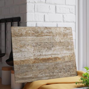 Può includere: Un tagliere rettangolare con un design a strati e strutturato nei toni del beige e del marrone. La tavola è appoggiata a un muro di mattoni bianchi, con un panno giallo sotto. Il design della tavola ricorda la pietra naturale o il legno.
