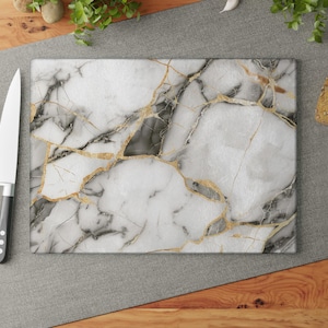 May include: Rectangular cutting board with a white marble design, featuring gold and grey veining. A stainless steel knife and red tomatoes are on the left. Bread, garlic, and herbs are in the background.