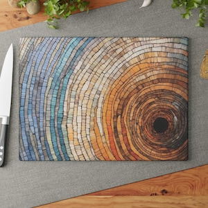 May include: Rectangular cutting board with a colorful, abstract design resembling tree rings. The design features shades of blue, orange, and brown. A large knife and tomatoes are on the left, with bread and garlic on the right.