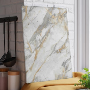 Peut inclure: Planche à découper rectangulaire en marbre avec veinage blanc, gris et doré. La planche est appuyée contre un mur de briques blanches. Des ustensiles de cuisine sont suspendus au mur à gauche. Un tissu jaune est en dessous.