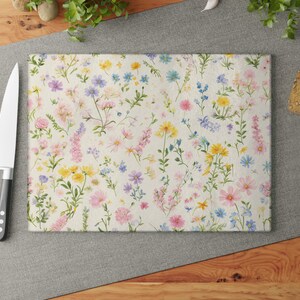 Puede incluir: Tabla de cortar rectangular con un estampado floral en colores pastel. El diseño presenta una variedad de flores rosas, amarillas, azules y moradas sobre un fondo crema. Un cuchillo de acero inoxidable y tomates rojos a la izquierda.