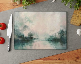 Puede incluir: Tabla de cortar rectangular de vidrio con una pintura de paisaje abstracto en tonos turquesa, verde y rosa. Un cuchillo de chef grande y dos tomates rojos están a la izquierda. La tabla está sobre una superficie gris.