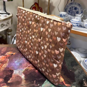 May include: A rectangular quilted pouch featuring a brown fabric base covered in a repeating pattern of small hearts in shades of pink, cream, and tan. The pouch has a top zipper closure and a small rectangular label reading "clms bag" on the front.