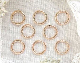 May include: Eight circular rose gold metal spring gate rings arranged in a grid pattern. Each ring features a small integrated spring-loaded mechanism that allows the ring to open and close securely for attaching various items or accessories.