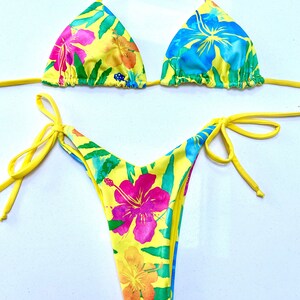 K&ouml;nnte beinhalten: Gelber Bikini mit tropischem Blumenmuster. Das Oberteil ist ein Dreieck-Modell mit verstellbaren Tr&auml;gern. Das Unterteil ist ein freches Modell mit B&auml;ndern an den Seiten.