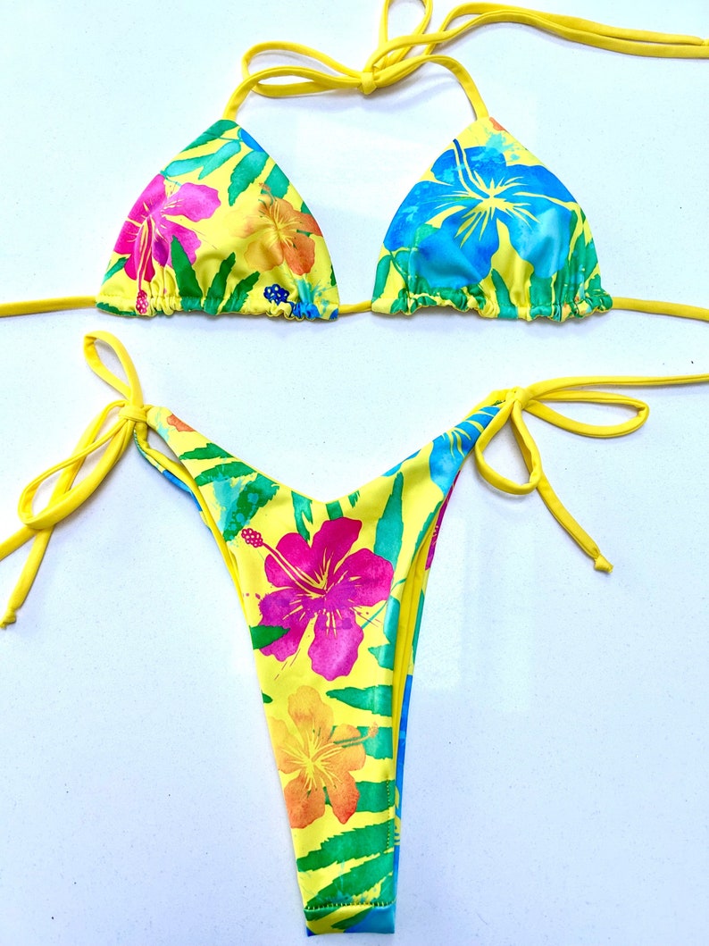 K&ouml;nnte beinhalten: Gelber Bikini mit tropischem Blumenmuster. Das Oberteil ist ein Dreieck-Modell mit verstellbaren Tr&auml;gern. Das Unterteil ist ein freches Modell mit B&auml;ndern an den Seiten.
