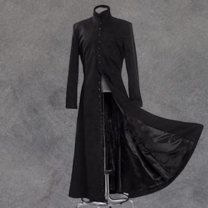 Puede incluir: Un abrigo largo y negro con cuello alto y abotonadura frontal. El abrigo está abierto, revelando un vistazo del forro y el interior de la prenda. El abrigo se exhibe en un maniquí sobre un fondo gris.