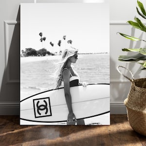 Puede incluir: Fotografía en blanco y negro de una mujer con una tabla de surf en la playa. La tabla de surf tiene un logotipo negro. La imagen se muestra en un lienzo contra una pared blanca. Una planta en una cesta tejida está a la derecha.