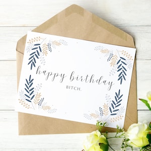 May include: A white greeting card with the text "happy birthday bitch" in black script, surrounded by blue, gold, and gray floral illustrations. The card is inside a brown envelope, with yellow roses in the foreground.
