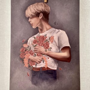 May include: A man with blond hair is holding a bouquet of red roses in front of his chest. He is wearing a white t-shirt with a floral pattern and blue jeans. The background is a muted gray.