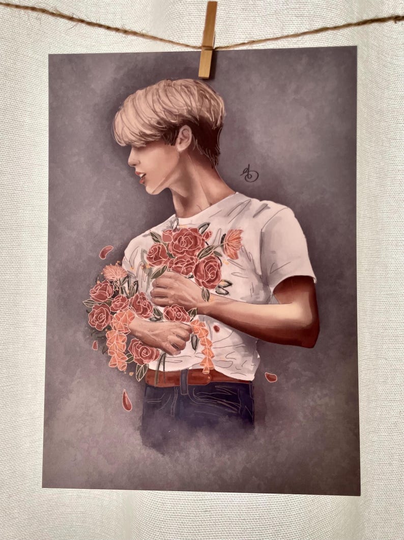 May include: A man with blond hair is holding a bouquet of red roses in front of his chest. He is wearing a white t-shirt with a floral pattern and blue jeans. The background is a muted gray.