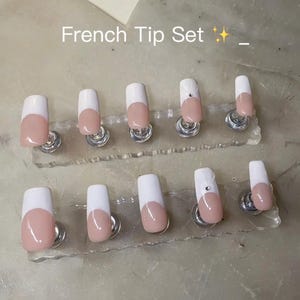 May include: A set of ten press-on nails with a French tip design, featuring a nude base and white tips. Two of the nails are accented with a small, clear rhinestone. The text "French Tip Set ✨ _" is visible above the nails on the display stands.