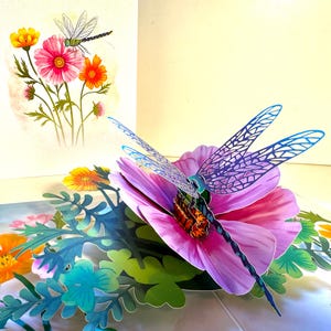 May include: A 3D pop-up greeting card with a dragonfly on a pink flower, with blue and purple details. The card also shows a watercolour illustration of flowers and a dragonfly. The theme is nature and floral.