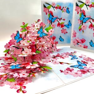 May include: A pop-up greeting card featuring a 3D floral arrangement of pink cherry blossoms and blue butterflies. The card is open, revealing layers of flowers and butterflies, with additional cards in the background.