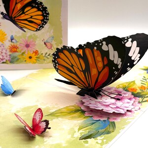 May include: A 3D pop-up greeting card featuring a monarch butterfly with orange, black, and white wings. The card has a watercolor floral design with pink, yellow, and blue flowers. A blue and red butterfly are also visible.