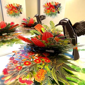 May include: Pop-up greeting cards featuring detailed illustrations of black birds with white markings, amidst vibrant, layered floral arrangements in red, orange, and green. The cards are displayed on a light-colored surface, creating a three-dimensional effect.