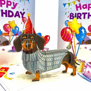 May include: A pop-up birthday card with a dachshund wearing a party hat and a grey sweater. The card has a "Happy Birthday" message, balloons, and a cake. The dog is brown, black, and tan.