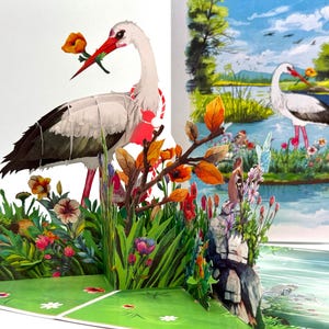 May include: A 3D pop-up greeting card featuring a white stork with black wingtips, holding a flower in its beak. The card has a vibrant, colorful design with flowers, foliage, and a painted landscape scene with a stork.