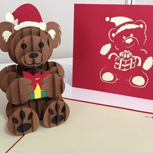 May include: A brown 3D pop-up Christmas card featuring a bear wearing a Santa hat and holding a gift. The card is on a cream-colored surface with a red border. A matching red card with a bear illustration is also present.