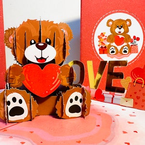Puede incluir: Una tarjeta pop-up 3D con un oso de peluche marrón que sostiene un corazón rojo. La tarjeta también tiene la palabra "LOVE" en letras doradas y una bolsa de regalo roja con corazones.