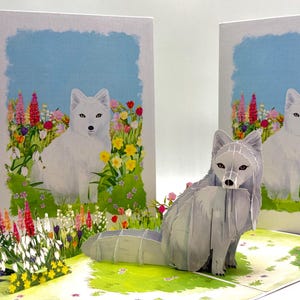 May include: A pop-up card with a white fox illustration in a flower field. A 3D grey fox model sits in front. The background shows a blue sky with a white border.