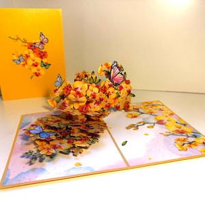 May include: A pop-up greeting card with a floral design, featuring orange and yellow flowers and butterflies. The card is open, displaying a 3D arrangement, with a yellow card in the background.