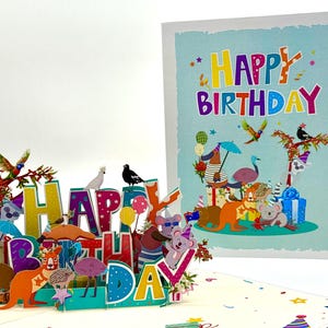 May include: A pop-up birthday card with a colorful design. The card features the words "HAPPY BIRTHDAY" in large, multi-colored letters, with various Australian animals and birds. A matching card is visible in the background.
