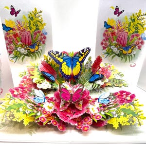 Puede incluir: Una tarjeta de felicitación desplegable con un vibrante arreglo floral. La tarjeta presenta flores rosas, amarillas y rojas, con varias mariposas coloridas con alas azules, amarillas y rosas.