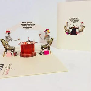 May include: A pop-up greeting card featuring a skeleton couple at a table with a bottle of wine. The card has the text "We are going to sit here and drink for as long as it takes to understand MEN!" The card is cream-colored.