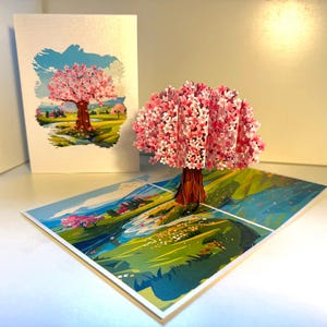 Puede incluir: Tarjeta de felicitación emergente 3D con un cerezo en flor rosa. La tarjeta abierta revela un paisaje detallado con un río y colinas. Una tarjeta a juego con una ilustración similar está detrás.