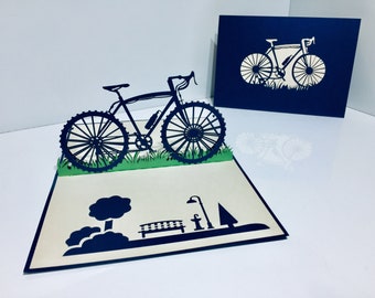 May include: A 3D pop-up card featuring a black silhouette of a bicycle with a bench, tree, and lamppost in the background. The card is open and the bicycle is the main focus of the image.