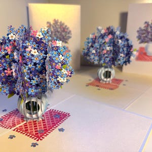 May include: Pop-up greeting cards featuring 3D floral arrangements. The cards display blue, pink, and white flowers in silver vases, set on red patterned mats. Additional cards with similar designs are visible in the background.