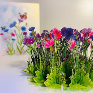 May include: A 3D pop-up card depicting a colourful meadow scene with pink, purple, and blue flowers. Butterflies are seen amongst the blooms. A matching flat card is visible in the background.