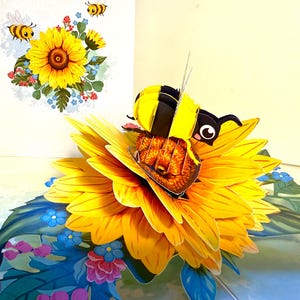 Puede incluir: Una tarjeta de felicitación emergente con un gran girasol amarillo con una abeja en 3D. La tarjeta tiene un fondo blanco con una imagen más pequeña de un girasol y dos abejas de dibujos animados. La abeja es negra y amarilla.
