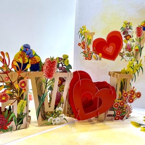 May include: A pop-up card with the word "LOVE" in a wooden font, adorned with vibrant flowers and two birds. The card showcases a large red heart and floral details, creating a lively and romantic design, perfect for a special occasion.