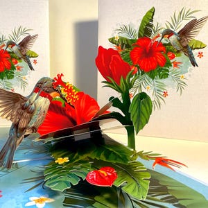 Puede incluir: Tarjeta de felicitación emergente 3D que presenta una escena tropical. Muestra un colibrí cerca de una gran flor de hibisco rojo, rodeado de exuberante follaje verde y otras flores coloridas. La tarjeta tiene un fondo azul claro.