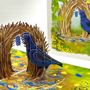 May include: A 3D pop-up card featuring a blue bird with a blue object in its beak, standing near a brown nest. The background shows a painted scene of a bird in a nest. The card is open, revealing the pop-up design.