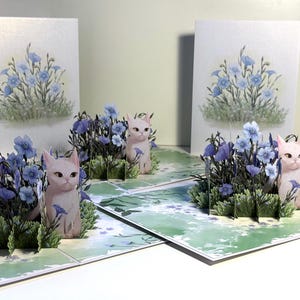 May include: Pop-up greeting cards featuring a garden scene with blue flowers and small pink cat figurines. The cards have a watercolor-style design with a light green and white background. The cards are folded open, displaying the 3D elements.
