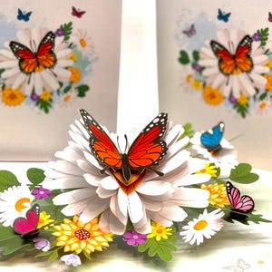 May include: A 3D pop-up greeting card featuring a large white daisy with a vibrant orange and black butterfly. Additional butterflies in blue and pink hover above the flower arrangement, with a background of other daisies and butterflies.