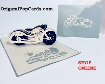 May include: A white and black 3D pop-up card featuring a motorcycle. The card is open and the motorcycle is in the center of the card. The card has a white background and the motorcycle is made of paper. The card is folded in half and the front of the card has the text "OrigamiPopCards.com" in black text. The back of the card has the text "SHOP ONLINE" in red text.