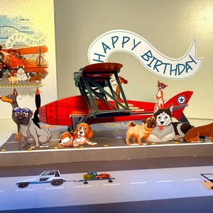 May include: A birthday card with a pop-up design showing a red biplane and several dog illustrations. The card includes a banner that reads "Happy Birthday." The background features an airport scene with an aeroplane.