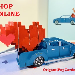 Puede incluir: Una camioneta azul con una caja llena de corazones pixelados rojos y rosas. La camioneta es una tarjeta pop-up con el texto "OrigamiPopCards.com" en la parte inferior.