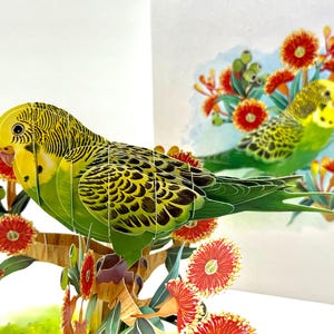 May include: A 3D pop-up greeting card featuring a green and yellow parakeet perched on a branch with red flowers. The bird has detailed feather patterns. The background shows a second parakeet and more flowers.
